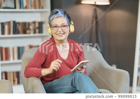 Senior woman is happily texting on her phone while wearing pink glasses and yellow headphones at home, reflecting a relaxed and modern lifestyle with a focus on technology and communication. High 99899813