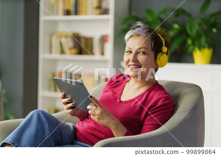 Mature woman smiles on a sofa, holding tablet PC and listening to music on headphones. She enjoys leisure, technology and music, feeling tranquil and blissful. High quality photo 99899864