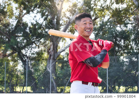 A young Asian man holding a baseball bat standing in a ballpark smiling A young Asian man holding a baseball bat standing in a ballpark smiling 99901804