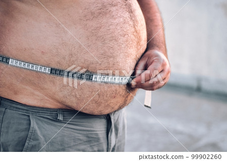 Close-up of a man's round and full belly, checking the width of his stomach with a measuring tape in centimeters 99902260