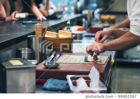 Close-up of a chef's hands rolling a sushi roll with rice and seaweed at a sushi bar with people in the background at the counter 99902267