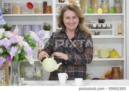 Beautiful happy ordinary elderly woman in the kitchen with a kettle and a cup Beautiful happy ordinary elderly woman in the kitchen with a kettle and a cup 99902895