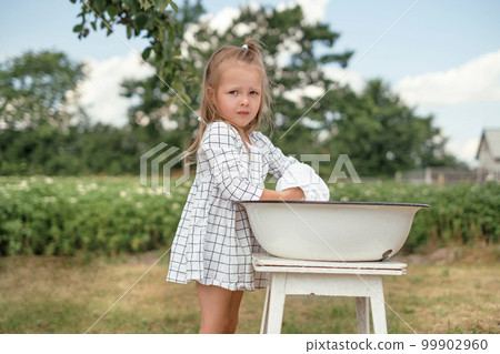 A beautiful attractive girl in a checkered dress is washing clothes in a basin in nature in the garden on a warm summer day. Little mom's assistant in household chores 99902960