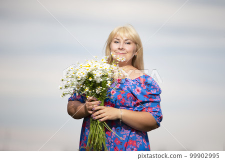 Beautiful plus size woman with white hair in a summer dress posing outdoors with daisies. Chubby girl in a meadow with flowers. 99902995