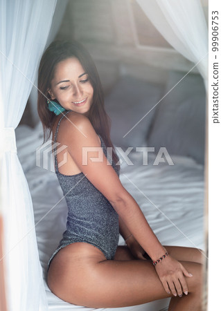 Beautiful tanned girl sitting posing on a white bed. The light on the right Beautiful tanned girl sitting posing on a white bed. The light on the right 99906753