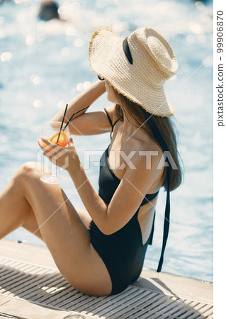 Young girl in a summer hat sitting near swimming pool. Girl wearing black swimwear and a hat. Girl posing for a photo near the pool and drinking a cocktail. 99906870