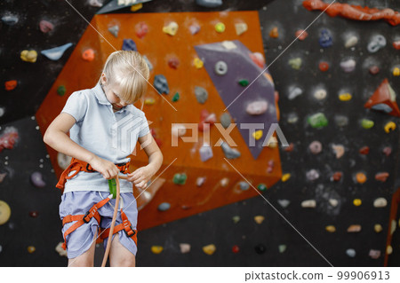 Little girl standing in rock-climbing gym. Girl is ready to climb up artificial wall wearing sports outfit and safety equipment. Blonde girl wearing shorts and t-shirt. Little girl standing in rock-climbing gym. Girl is ready to climb up artificial wall wearing sports outfit and safety equipment. Blonde girl wearing shorts and t-shirt. 99906913