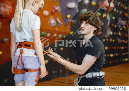 Instructor man is helping client small girl to put on safety harnesses in rock climbing center indoors. Little girl is engaged in rock climbing in the children's entertainment center. Blonde caucasian 99906918