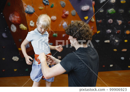 Instructor man is helping client small girl to put on safety harnesses in rock climbing center indoors. Little girl is engaged in rock climbing in the children's entertainment center. Blonde caucasian 99906923