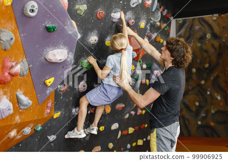 Male instructor is helping a little girl in climbing gym to climb on artificial rocks on wall. Little girl is engaged in rock climbing in the children's entertainment center. Blonde caucasian girl 99906925