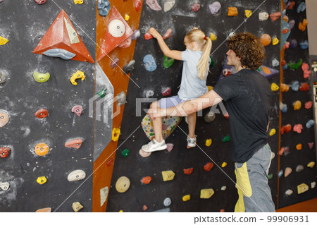 Male instructor is helping a little girl in climbing gym to climb on artificial rocks on wall. Little girl is engaged in rock climbing in the children's entertainment center. Blonde caucasian girl 99906931