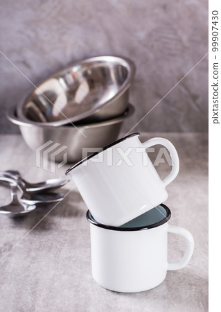 Two white empty metal mugs and metal bowls and spoons on a gray vertical background 99907430