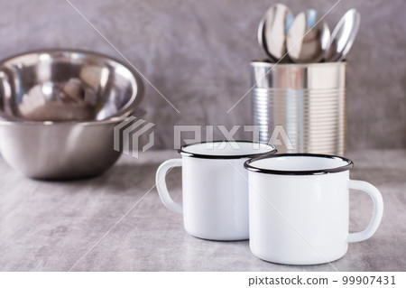 Two white empty metal mugs, spoons in a can and metal plates on a gray background 99907431