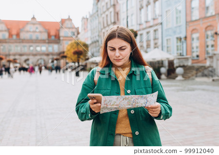 Sad young female tourist is exploring new city. Redhead girl walking in old town and holding a paper map and smart phone in a sunny day. Urban lifestyle concept Sad young female tourist is exploring new city. Redhead girl walking in old town and holding a paper map and smart phone in a sunny day. Urban lifestyle concept 99912040