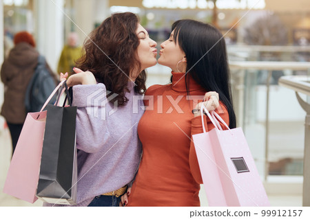 Horizontal shot of attractive happy young women with shopping bags enjoying shopping, standing and kiss each other, ladies wearing casual sweaters, buying new clothing. Shopping and lifestyle concept. 99912157