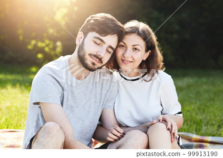 Romantic couple sitting close to each other while resting outdoor at nature during summer day, feeling happiness and relaxation. People, love, relationships, harmony and togetherness concept 99913190