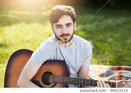 Young bearded male guitarist in casual T-shirt sitting with musical instrument on green grass, playing different songs, looking at camera with serious look, resting at nature. People, hobby, music 99913218