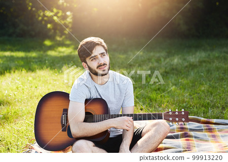 Outdoor portrait of attractive young man with beard dressed casually while resting on green grass with guitar, having pensive expression, dreaming about something in his life. Romantic man on nature 99913220