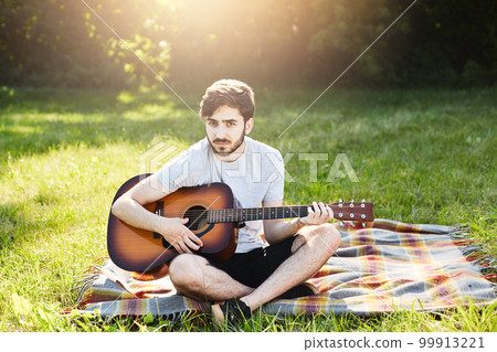 Handsome bearded man wearing summer clothes sitting on coverlet with guitar, enjoying sunny weather and beautiful landscapes, keeping his legs crossed, feeling enjoyment and relaxation. Male guitarist 99913221