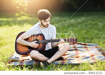 Portrait of attractive stylish bearded guy with guitar sitting crossed legs on green grass, playing musical instrument, enjoying calm atmosphere. Talented boy demonstrating his talent while resting Portrait of attractive stylish bearded guy with guitar sitting crossed legs on green grass, playing musical instrument, enjoying calm atmosphere. Talented boy demonstrating his talent while resting 99913222