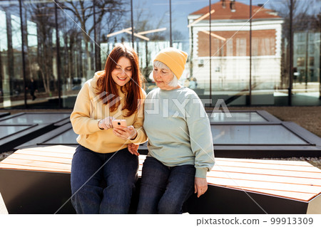 Two women together wear casual clothes using smart phone and sitting on the bench on city street. Family day concept. Young woman and senior middle aged mom spending time together 99913309