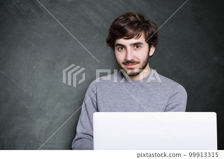 A confident attractive guy with dark beard and stylish hairstyle sitting in front of open laptop computer having charming smile looking into camera with his big brown eyes. A confident attractive guy with dark beard and stylish hairstyle sitting in front of open laptop computer having charming smile looking into camera with his big brown eyes. 99913315