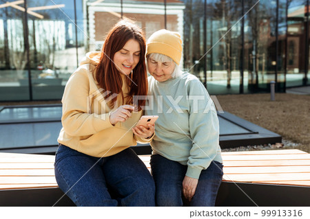 Two women together wear casual clothes using smart phone and sitting on the bench on city street. Family day concept. Young woman and senior middle aged mom spending time together Two women together wear casual clothes using smart phone and sitting on the bench on city street. Family day concept. Young woman and senior middle aged mom spending time together 99913316