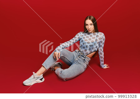 Young girl in transparent top and jeans sits on the floor in red background Young girl in transparent top and jeans sits on the floor in red background 99913328