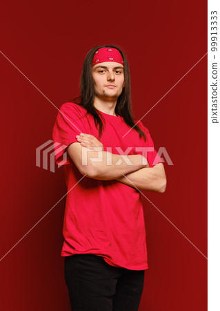 A young adult man stands in a studio with arms crossed and looks confidently at the camera against red background 99913333
