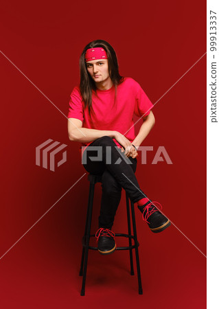 Young man sits on high stool with his leg on another leg and leaning forward Young man sits on high stool with his leg on another leg and leaning forward 99913337