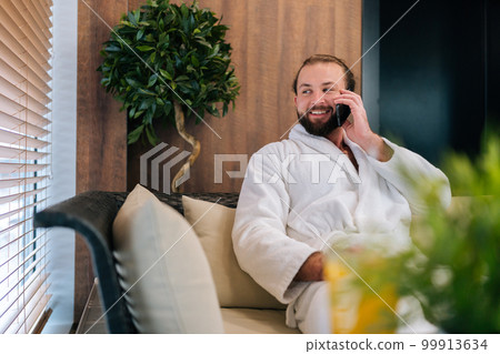 Portrait of joyful bearded young man wearing white bathrobe talking using smartphone smiling looking away, sitting by window on comfy sofa at spa salon after beauty procedures. 99913634