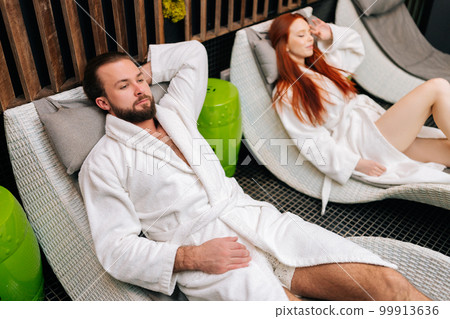 High-angle view of young couple in white bathrobes lying on comfy loungers by poolside after spa treatment at beauty salon. Bearded man and pretty woman enjoying relaxing after body massage treatment High-angle view of young couple in white bathrobes lying on comfy loungers by poolside after spa treatment at beauty salon. Bearded man and pretty woman enjoying relaxing after body massage treatment 99913636