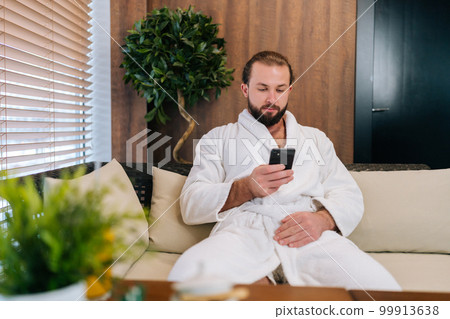 Portrait of handsome young man with beard wearing white bathrobe using mobile phone looking to device screen, sitting by window on comfy sofa at spa salon after beauty procedures. Portrait of handsome young man with beard wearing white bathrobe using mobile phone looking to device screen, sitting by window on comfy sofa at spa salon after beauty procedures. 99913638