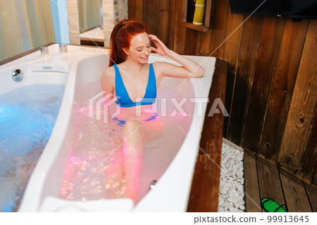 High-angle view of happy young woman relaxed lying in bath with hydromassage at luxury spa salon. Pretty redhead female sitting in white bathtub and relaxing hydrotherapy in beauty center. High-angle view of happy young woman relaxed lying in bath with hydromassage at luxury spa salon. Pretty redhead female sitting in white bathtub and relaxing hydrotherapy in beauty center. 99913645