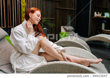 Side view of cheerful young woman in white bathrobe relaxing sitting on deck chair by poolside with closed eyes after beauty treatment at spa salon. Pretty female enjoying wellness weekend. Side view of cheerful young woman in white bathrobe relaxing sitting on deck chair by poolside with closed eyes after beauty treatment at spa salon. Pretty female enjoying wellness weekend. 99913647