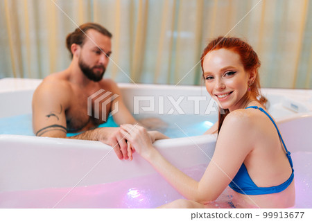Portrait of cheerful young woman in bikini and brutal bearded man sitting in double hot bath tub with hydromassage holding hands at luxury spa salon, enjoying weekend in bathtub. Portrait of cheerful young woman in bikini and brutal bearded man sitting in double hot bath tub with hydromassage holding hands at luxury spa salon, enjoying weekend in bathtub. 99913677