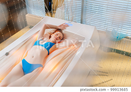 High-angle view of carefree young woman in bikini lying with closed eyes on electric massage bed, having rest enjoying spa procedure at luxury spa salon. Sensual redhead female enjoying wellness. 99913685