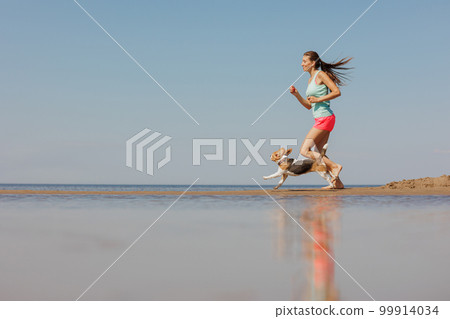 athletic woman and beagle dog run along sandy beach on seashore in morning. outdoor 99914034