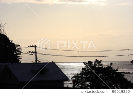 淺橙色的天空、雲彩、剪影的房屋、樹木、電線、大海和光亮的海面上的地平線 淺橙色的天空、雲彩、剪影的房屋、樹木、電線、大海和光亮的海面上的地平線 99919230