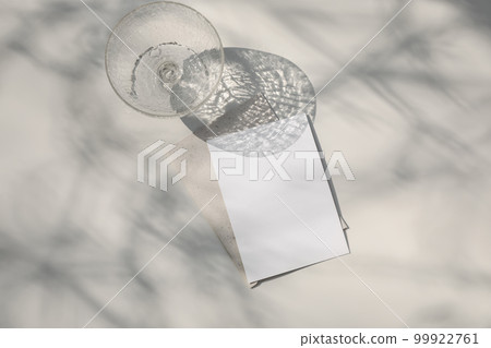 Trendy summer stationery still life. Champagne, cocktail glass with long shadows. Blank greeting card, invitation mock up. Beige table background in sunlight. Tree branches shadows overlay. Flat lay. Trendy summer stationery still life. Champagne, cocktail glass with long shadows. Blank greeting card, invitation mock up. Beige table background in sunlight. Tree branches shadows overlay. Flat lay. 99922761