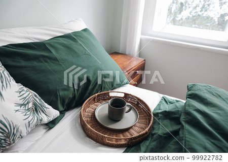 Winter breakfast in bed composition. Cup of coffee on wicker tray. Dark green linen bedding. Lifestyle composition. Scandinavian bedroom, interior. Blurred window in background. Top view. Winter breakfast in bed composition. Cup of coffee on wicker tray. Dark green linen bedding. Lifestyle composition. Scandinavian bedroom, interior. Blurred window in background. Top view. 99922782