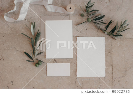 Italian summer wedding stationery set. Mock-up scene with blank paper greeting, RSVP cards on beige marble tiles background. Green olive tree branches, silk ribbon. Mediterranean flat lay, top view. 99922785