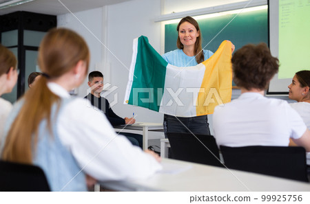 High school teacher tells students about Ireland and holds a Ireland flag in hands. 99925756