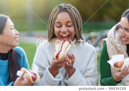 Friends eating hot dogs in a park 99934189
