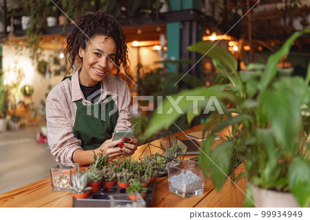 Smiling woman florist wearing apron holding phone during working day in floral shop Smiling woman florist wearing apron holding phone during working day in floral shop 99934949