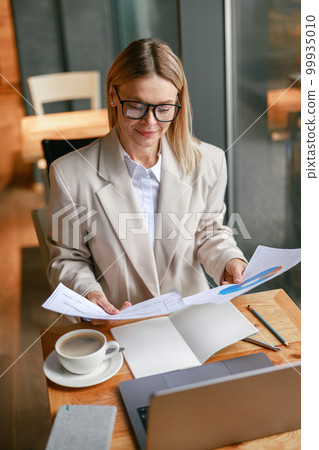 Smart businesswoman in glasses working with documents and use laptop while sitting in cafe Smart businesswoman in glasses working with documents and use laptop while sitting in cafe 99935010