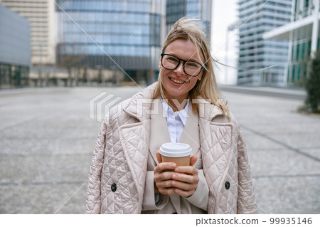Attractive businesswoman holding paper cup of coffee while standing on business center background Attractive businesswoman holding paper cup of coffee while standing on business center background 99935146