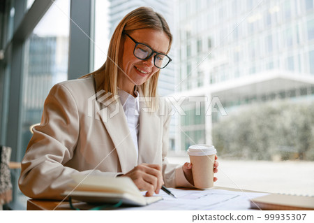 Smiling businesswoman in glasses drinking coffee and making notes while working in cafe Smiling businesswoman in glasses drinking coffee and making notes while working in cafe 99935307