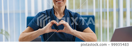 Smiling doctor working laptop and showing heart with hands in medical office Smiling doctor working laptop and showing heart with hands in medical office 99935483