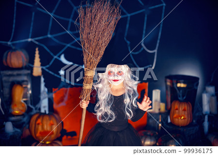 Kid girl in witch costume scaring, making faces with broom in hand on background decor Halloween 99936175